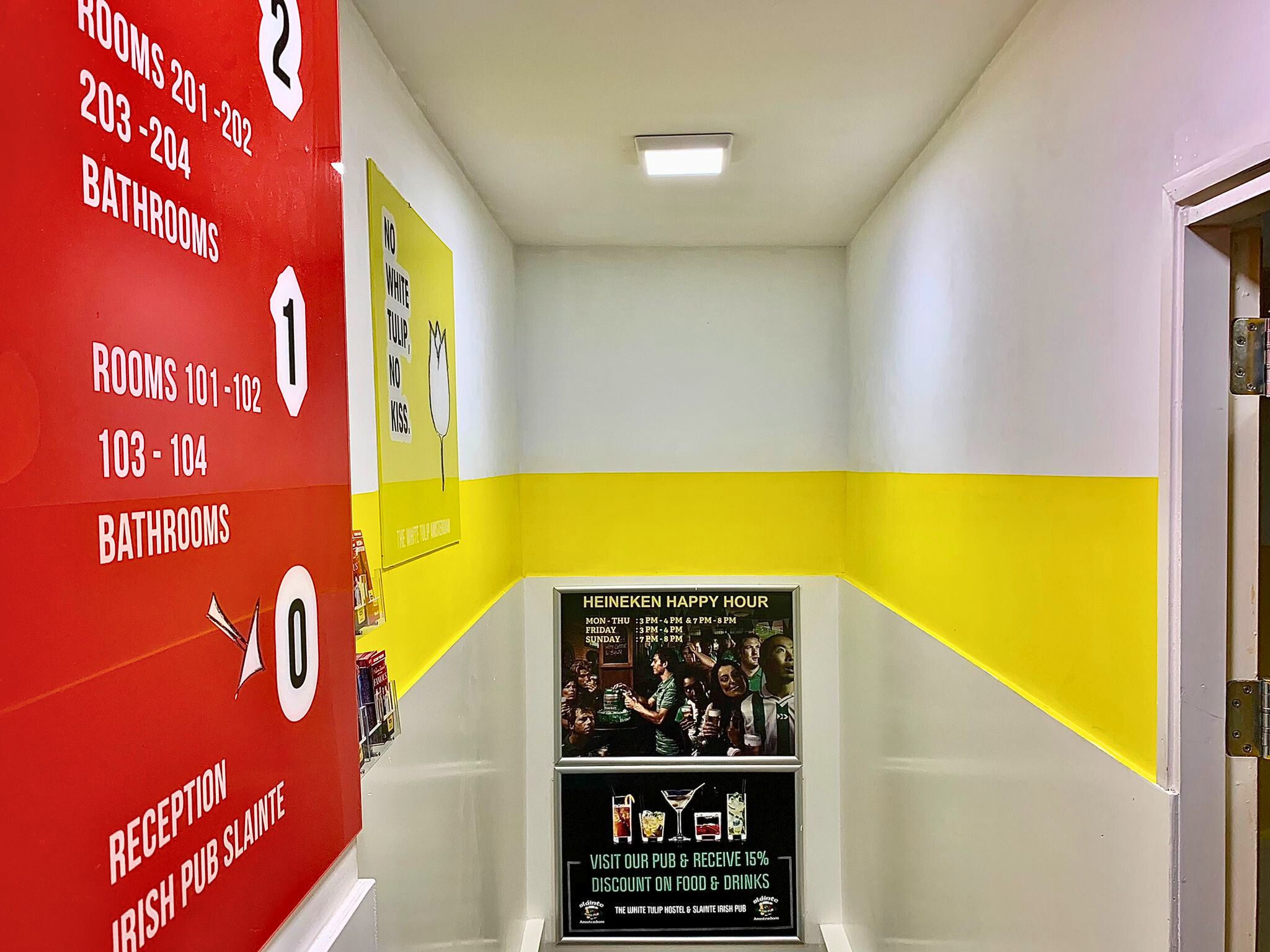 A brightly colored corridor with directional signage. To the left, a red wall with white text indicating room numbers and facilities on different floors; ahead, a poster for Heineken Happy Hour; and to the right, a yellow wall with a playful text poster.