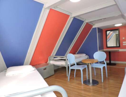 A colorful attic room with slanted blue and red walls, featuring a single bed, a table with two blue chairs, a heater, and a wall-mounted flat screen TV.