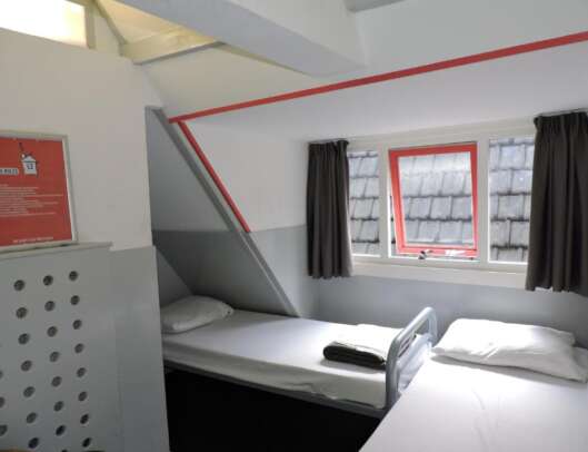 A small, minimalist dormitory room with a pitched roof, a red-framed window with grey curtains, two white beds with pillows, and a list of house rules on the wall.