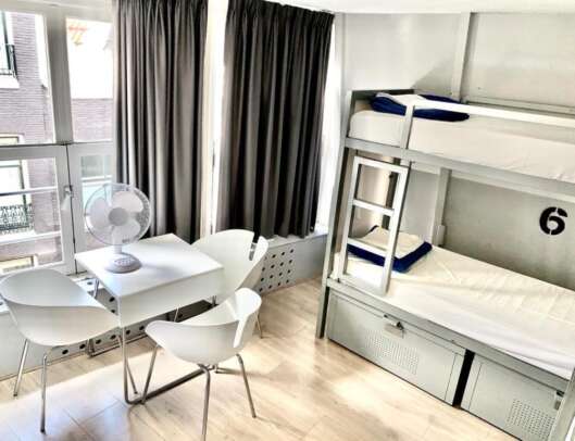 A bright, modern room with a white bunk bed set against a wall with a number 6 decal, a dining area with a white table and four white chairs, a white standing fan on the table, dark curtains on a window that offers a view of building facades, and light wooden flooring.
