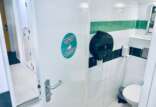 A modern public restroom with a half-open door revealing a white toilet and a black toilet paper dispenser on a white and green tiled wall, with a soap dispenser on the right; a sticker on the door reads, "We Aim to Please So Please Aim."