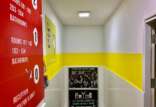 A brightly-lit corner in a hallway with walls painted in yellow and red and directional signs indicating room numbers, bathrooms, and reception, with an advertisement for a Heineken Happy Hour on the wall.