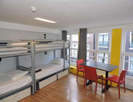 A hostel room with two metal bunk beds and a window overlooking a street, featuring a wooden floor, a small dining table with red chairs, and yellow storage lockers.