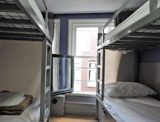A dormitory room with two sets of metal bunk beds, clean bedding, a window providing natural light, and neutral colored walls.