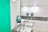 A modern public bathroom interior with a bright green accent wall, featuring two white sinks with silver faucets, accompanied by two wall-mounted soap dispensers and a large mirror overhead.
