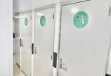 Three white restroom doors in a corridor, each marked with green circular signs indicating gender-neutral toilets, with one displaying a symbol for disability access.