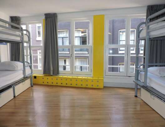 A hostel dormitory room with two metal bunk beds, hardwood flooring, a large window with grey curtains, and a yellow-painted central heating column.