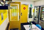 A small, brightly lit break room with yellow doors, a white table with a vase of tulips, blue chairs, and two vending machines against the far wall. There are informational posters on the wall and a bench with a shelf holding books and magazines above it.