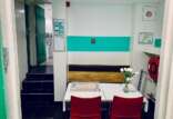 A small break room with a white table, red chairs, a wooden bench with a turquoise stripe on the wall, and a vase with white tulips. To the left is a kitchen area, and fire safety equipment is mounted on the walls.