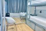 A hostel dormitory room with a metal bunk bed, the lower bunk is numbered 4, white chairs, a small table, curtains, and a trash bin.